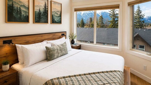 A bedroom with a large bed, a nightstand, a window with a mountain view, and three framed pictures on the wall. - "Alpine Hideaway" at Alpine Village (Canmore)