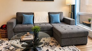 A modern living room with a grey sectional sofa, a glass coffee table with books and a plant, a painting of a mountain landscape, and a lamp by the window. - "Alpine Hideaway" at Alpine Village (Canmore)
