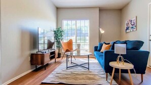 Ein Wohnzimmer mit einem blauen Sofa, einem kleinen runden Tisch, einem Sessel, einer Stehlampe, einer Pflanze und einem Fernseher.