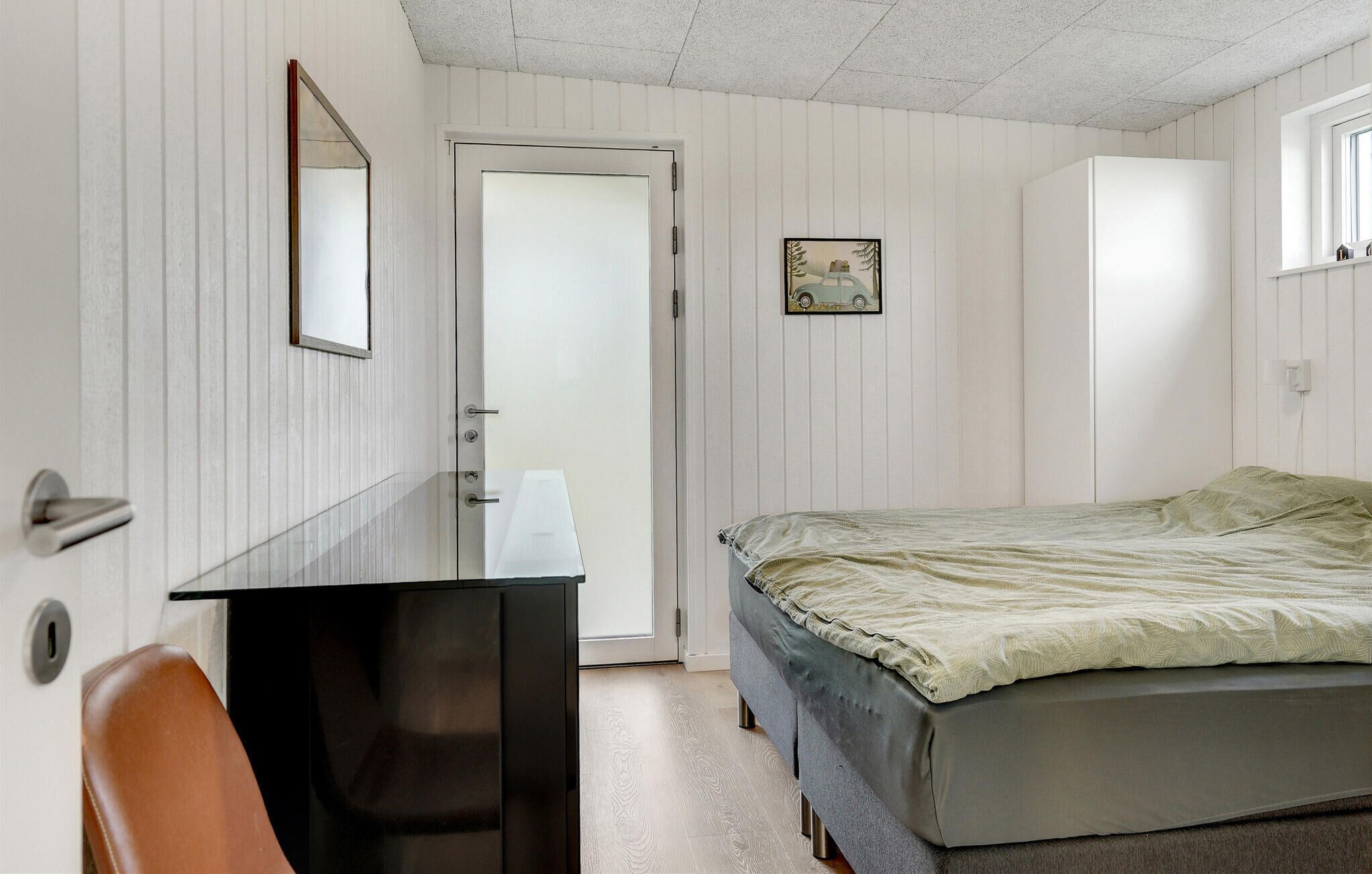 Une chambre à coucher avec un lit, un bureau, une chaise, une porte et un petit tableau accroché au mur.