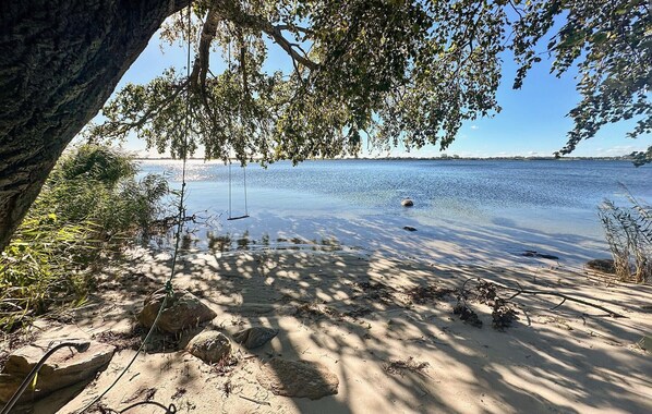 Une plage de sable avec une aire de jeux comprenant des balançoires, des arbres et une étendue d’eau calme.