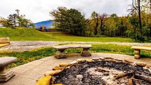 A stone fire pit surrounded by a stone bench and a stone table, set in a grassy area with trees and a hill in the background. - 2-bedroom condo in fantastic Gatlinburg with AC (Gatlinburg)