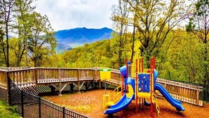 A playground with a slide and climbing structures, surrounded by a wooden fence and trees, with mountains in the background. - 2-bedroom condo in fantastic Gatlinburg with AC (Gatlinburg)