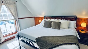 Un dormitorio con cama, almohadas, lámpara de noche, ventana con cortinas y vista al exterior.