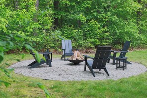 A circular outdoor seating area with four Adirondack chairs and a central fire pit surrounded by gravel.