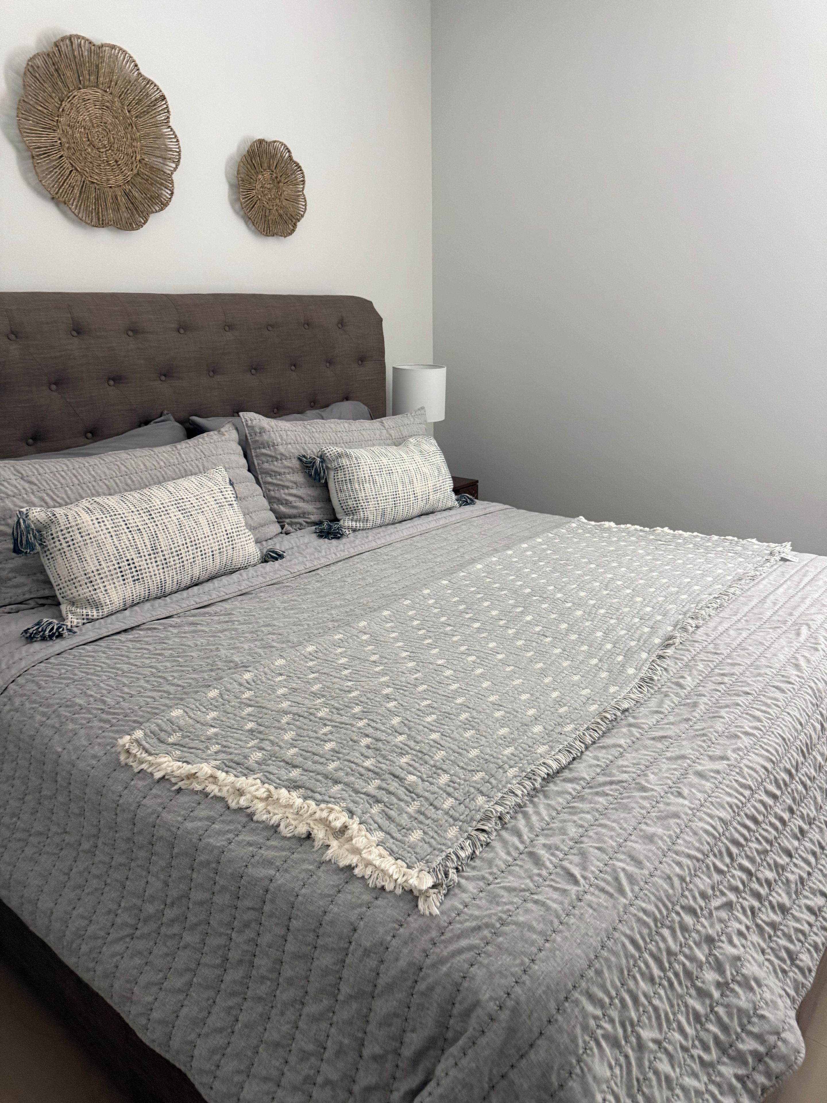 A neatly made bed with a grey quilt and pillows, a tufted headboard, and two wall-mounted decorative items.