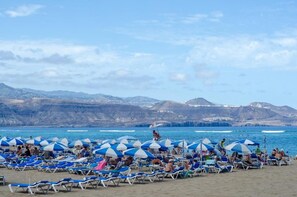 A beach with rows of sunbeds and umbrellas, a clear sky, and mountains in the background. - Home2Book Bright Penthouse with Terrace & Views (Las Palmas de Gran Canaria - Cuenca del Guiniguada)