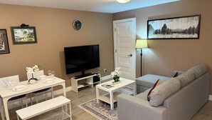 Een woonkamer met een flatscreen televisie, een bank, een eettafel met stoelen, een bankje, een klok en een schilderij aan de muur.
