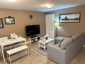 Ein Wohnzimmer mit Fernseher, einer Couch, einem Esstisch mit Stühlen, einer Bank, einer Uhr und einem Gemälde an der Wand.