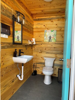 A wooden-paneled bathroom with a white sink, black faucet, and a toilet. There are two wall-mounted light fixtures and a painting above the toilet. - Dale-Vintage Glamping at Winery Near Sequoia (Dunlap)