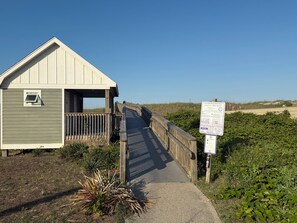 A wooden boardwalk leading to a small building with a sloped roof, surrounded by vegetation and a clear sky.