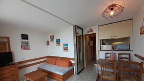 Un ambiente compatto con divano, tavolo in legno, sedie, televisore e angolo cottura.