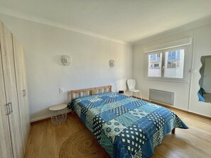 Une chambre à coucher comprenant un lit, une chaise, une petite table et une fenêtre donnant sur des immeubles.