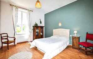 Une chambre à coucher comprenant un lit, une chaise, une petite table, une lampe, une armoire et une fenêtre avec des rideaux.