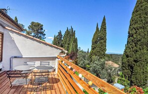 Une terrasse en bois avec des meubles d’extérieur, un bâtiment blanc doté d’un toit en pente, et de grands cyprès en arrière-plan.
