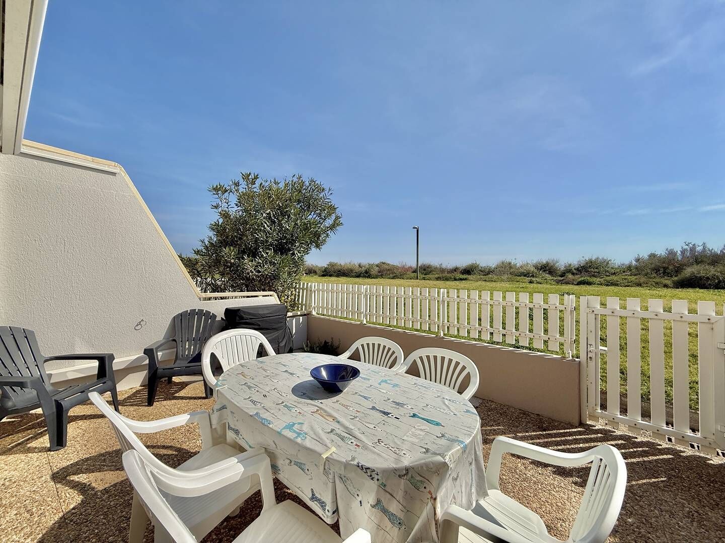 Een terras buiten met een tafel en stoelen, een wit hek en uitzicht op een groen veld.