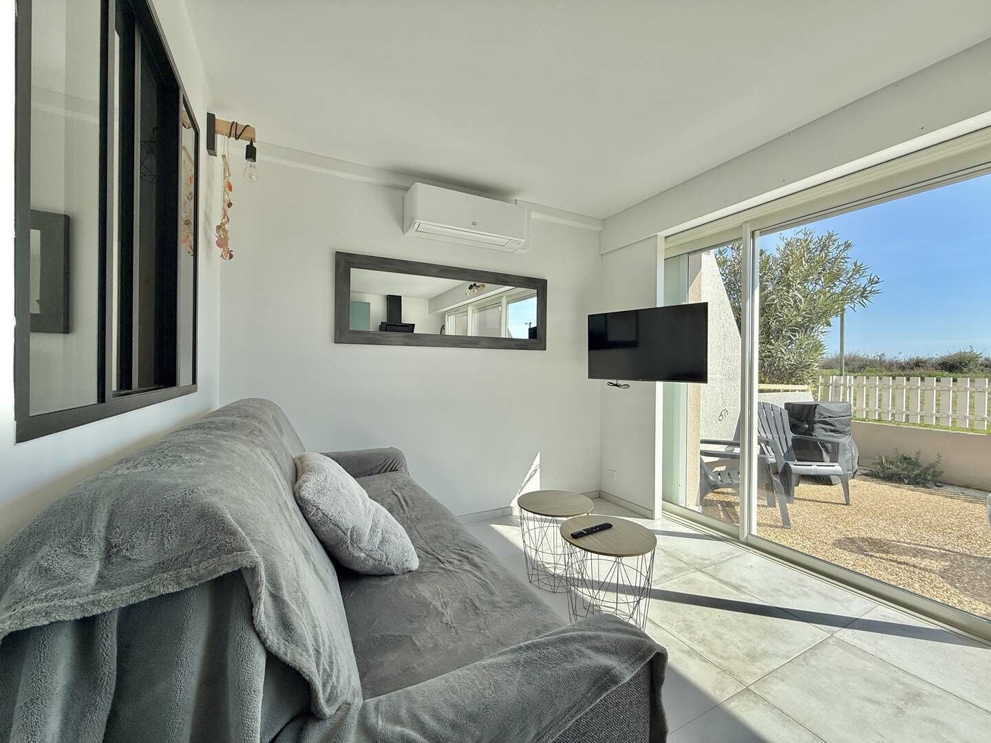Una sala de estar moderna con un sofá gris, un televisor de pantalla plana y un ventanal corredero que da a un espacio exterior con una silla de patio.