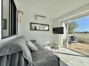 A modern living room with a grey sofa, a flat-screen TV, and a sliding glass door leading to an outdoor area with a patio chair. - CAP D'AGDE - Plage Richelieu / Réf 0304 (CAP D'AGDE)