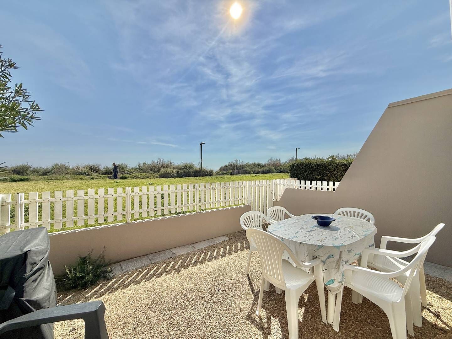 An outdoor patio with a table and chairs, a white fence, and a view of a grassy field.