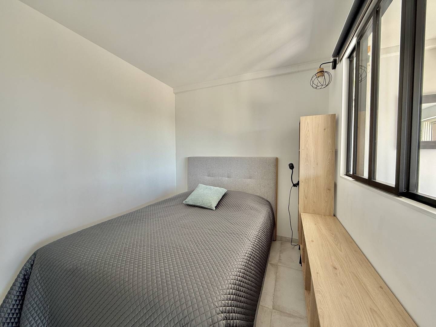 A single bed with a grey quilted bedspread and a light-colored pillow, a wooden bench, and a window with a metal frame.