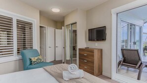 Een slaapkamer met een bed, een stoel, een dressoir, een televisie en een schuifdeur naar een balkon.
