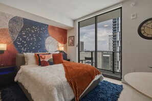 Un dormitorio moderno con una cama grande, vista a la ciudad a través de una puerta corrediza, un reloj de pared y un cuadro.
