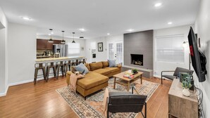 Una sala moderna con sofá, mesa de centro y televisor. Se observa un área de cocina con taburetes y una chimenea.