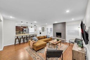Ein modernes Wohnzimmer mit Sofa, Couchtisch und Fernseher. Es gibt eine Küchenzeile mit Barhocker und einen Kamin.