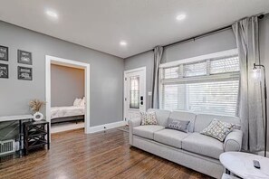 A modern living room with a grey sofa, a small table, a bed in the background, and a large window with blinds. - Fully renovated in 2023 to provide modern amenities in a century home. (Cleveland)