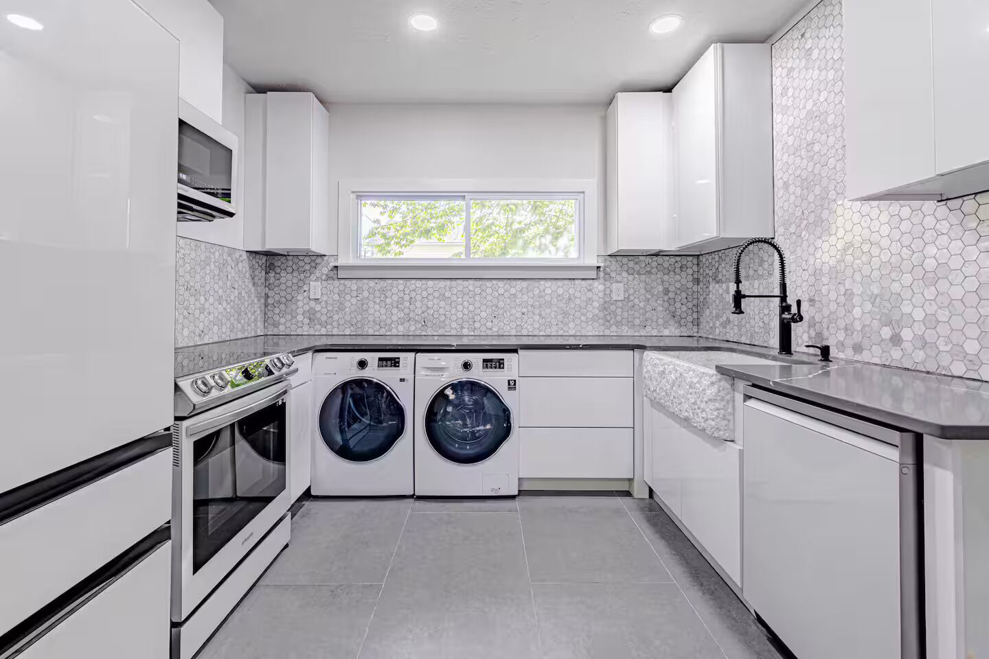 A modern kitchen with a built-in oven, two wall-mounted washing machines, a window, and a tiled backsplash.