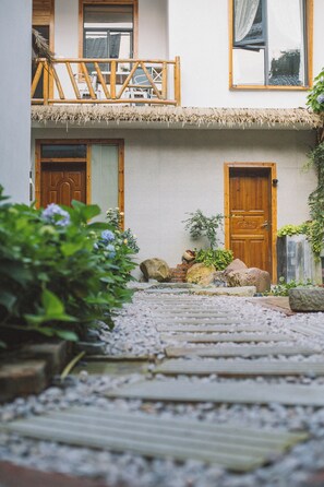 A building with a wooden door, a balcony with a thatched roof, and a gravel pathway leading to the entrance.