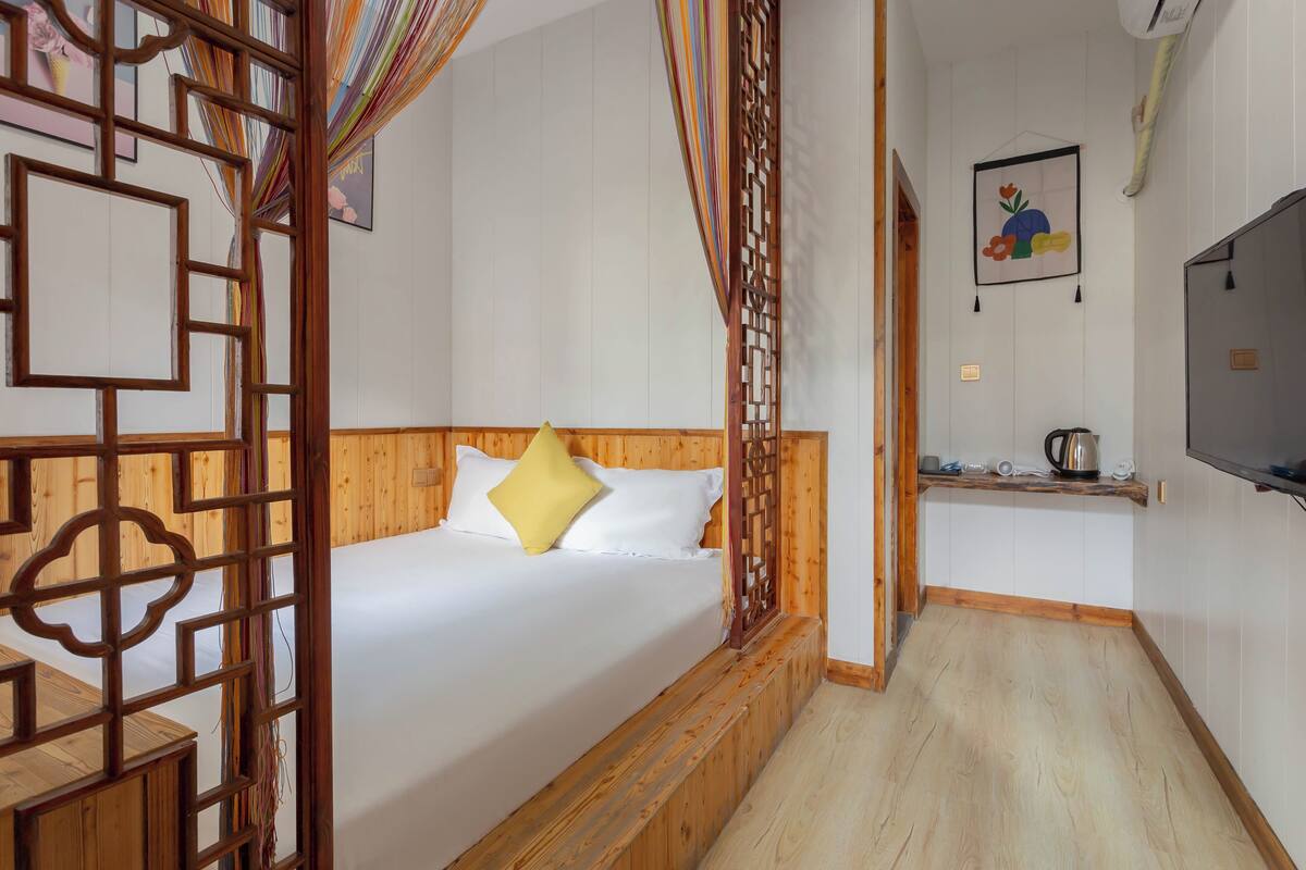 A bedroom with a wooden headboard, a bed with white bedding and a yellow pillow, a TV on the wall, and a small kitchenette area.