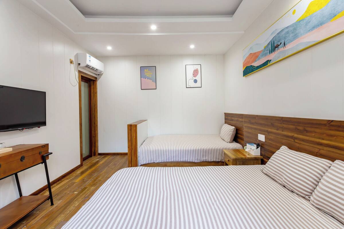 A hotel room with a large bed, a smaller bed, a desk with a TV, a wooden floor, and a painting on the wall.