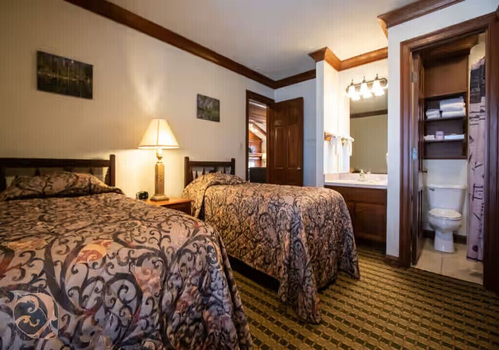 Une chambre d’hôtel avec deux lits, une lampe de chevet, une salle de bain avec des toilettes et un lavabo, et un placard avec des étagères.