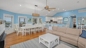 Un vaste espace de vie comprenant une table à manger, des chaises blanches, un canapé beige et un ventilateur de plafond.
