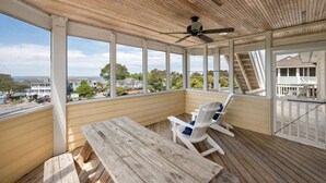 Un espace extérieur abrité, doté d’une table et de chaises en bois, et de grandes fenêtres offrant une vue sur l’océan et les bâtiments environnants.