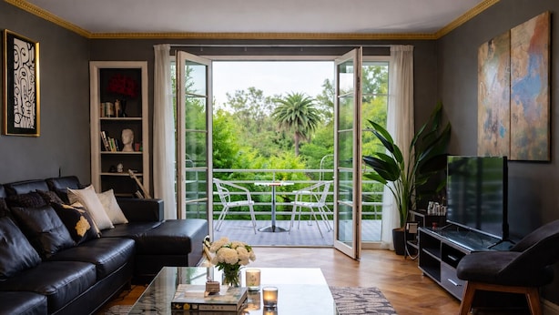 Een woonkamer met een zwarte leren bank, een glazen salontafel, een boekenkast, een televisie en een balkon met uitzicht op groen.