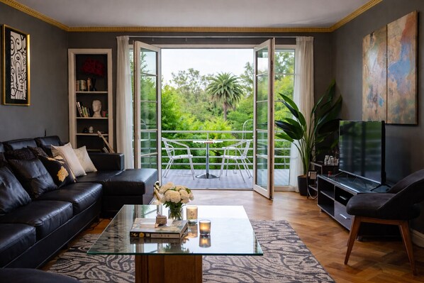 A living room with a black leather sofa, a glass coffee table, a bookshelf, a TV, and a balcony with a view of greenery. - Spacious patrimonial apartment for 7 people. View of the park. Metro Bellas Artes. (Santiago)