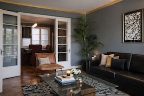 A modern living room with a glass coffee table, leather sofa, and a dining area visible through an open door. - Spacious patrimonial apartment for 7 people. View of the park. Metro Bellas Artes. (Santiago)