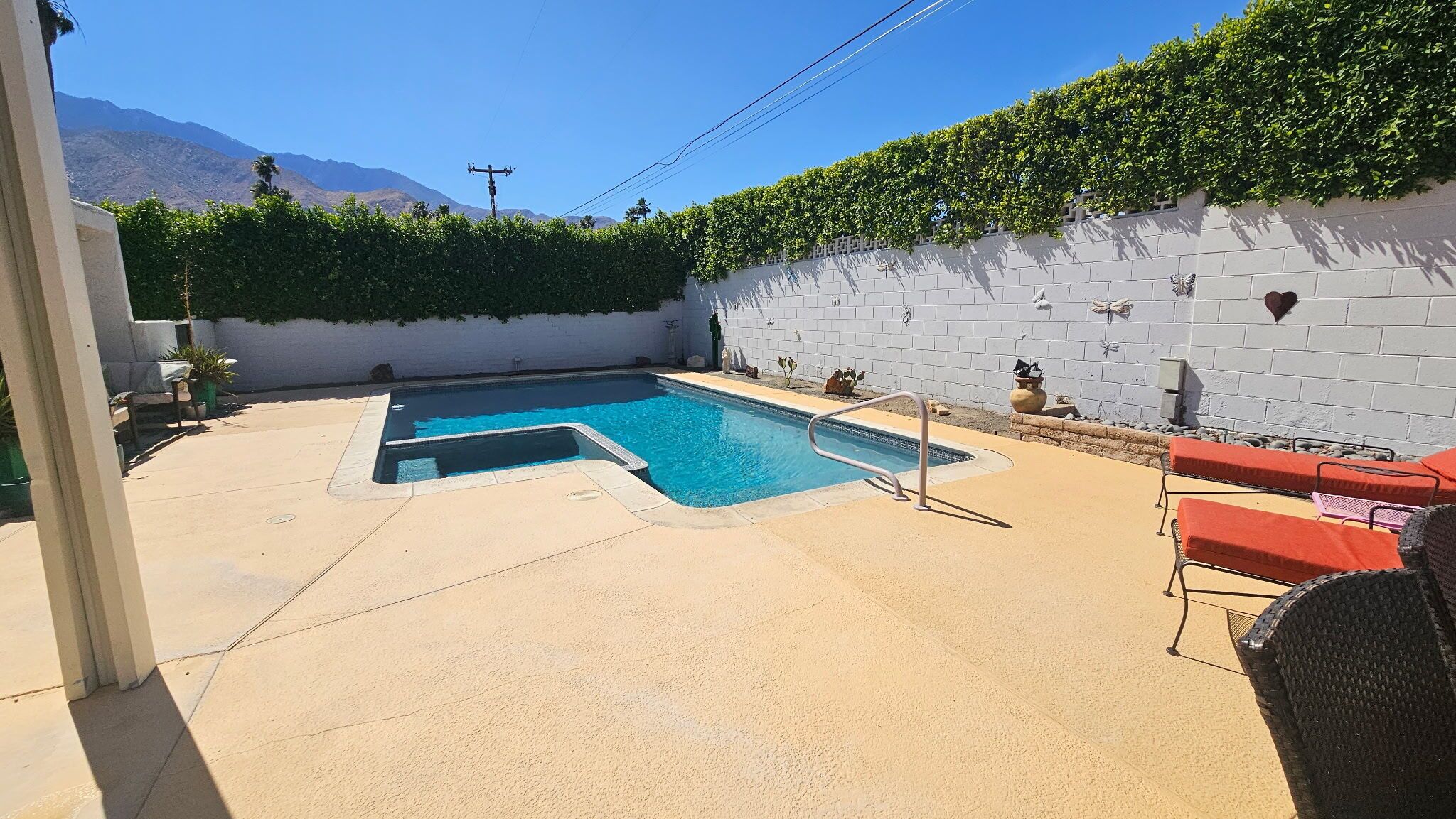 Piscina con pared blanca y seto, rodeada de un patio de hormigón y mobiliario de exterior.