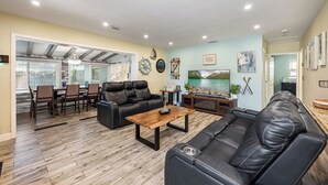 A spacious living room with a large black leather sofa, a wooden coffee table, and a TV on a wooden stand. There is a dining area with a table and chairs, and a kitchen area visible through an open door. - Family 3BR w/ Pool | Game Room I Walk to WaterPark (Margate)