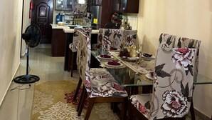 A dining area with a glass table, floral chairs, and a kitchen in the background.