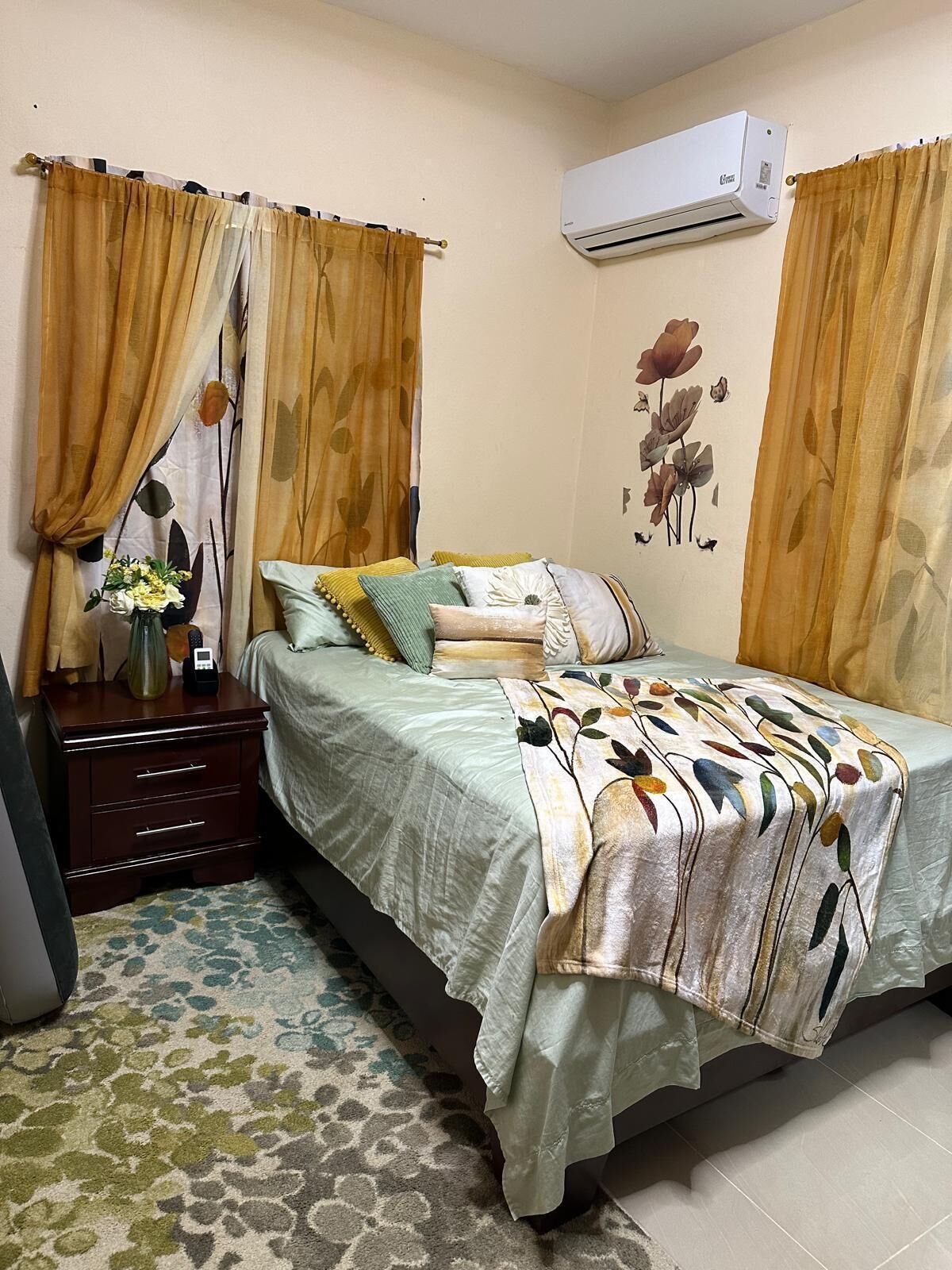 Un dormitorio con cama, mesita de noche, aire acondicionado y decoración floral en la pared.