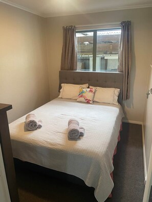 Een net opgemaakt bed met een witte dekbedovertrek en twee opgerolde handdoeken, een raam met gordijnen en uitzicht op een gebouw.
