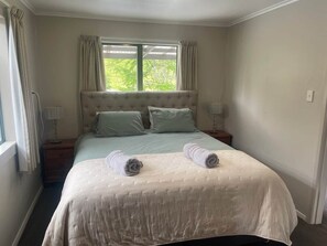 A neatly made bed with two rolled towels, a window with curtains, and a bedside table with a lamp. - Thompson Country Cottage (Kapenga)