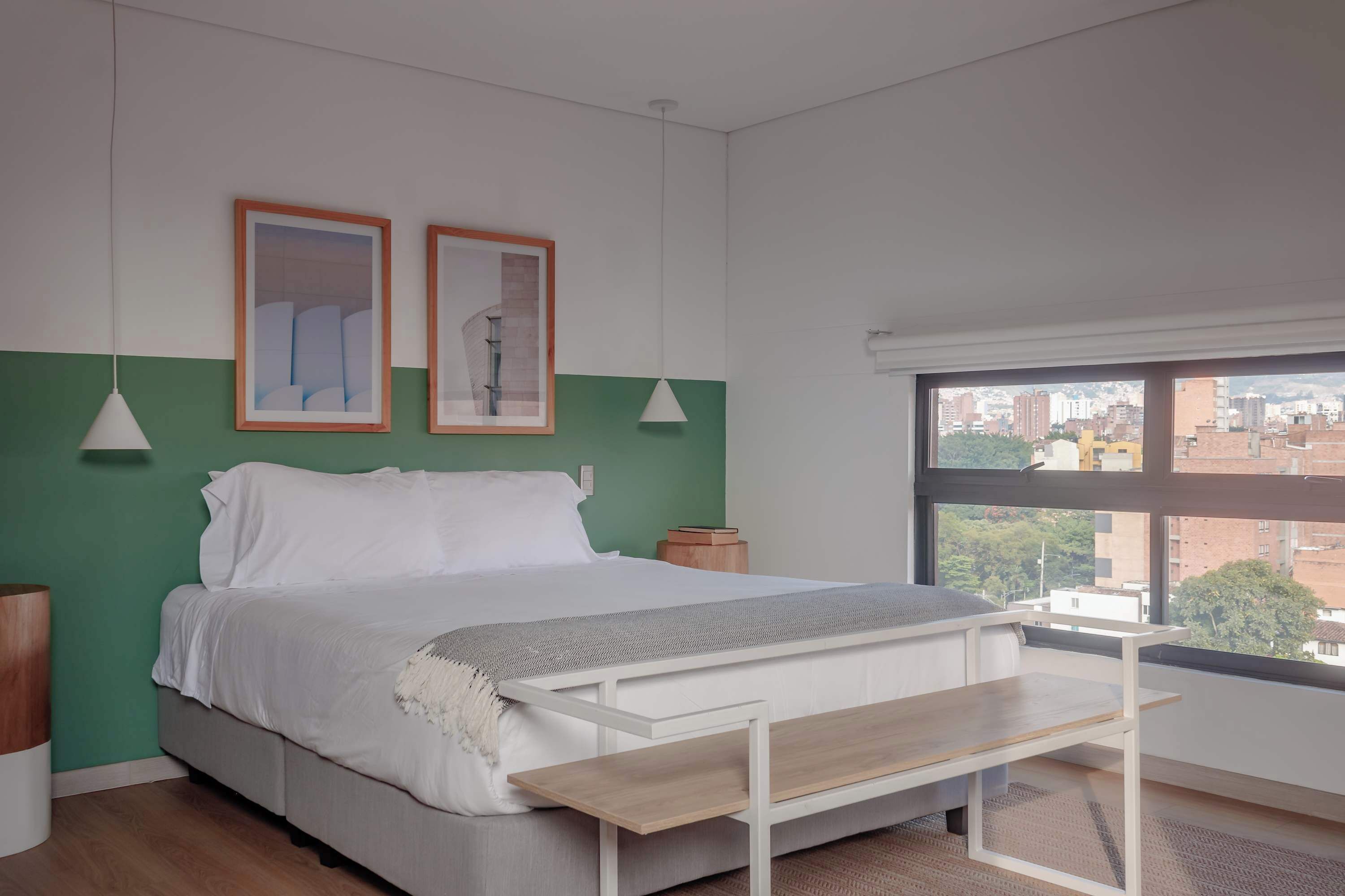 Een slaapkamer met een bed, nachtkastje, raam met uitzicht op de stad en twee ingelijste foto's aan de muur.