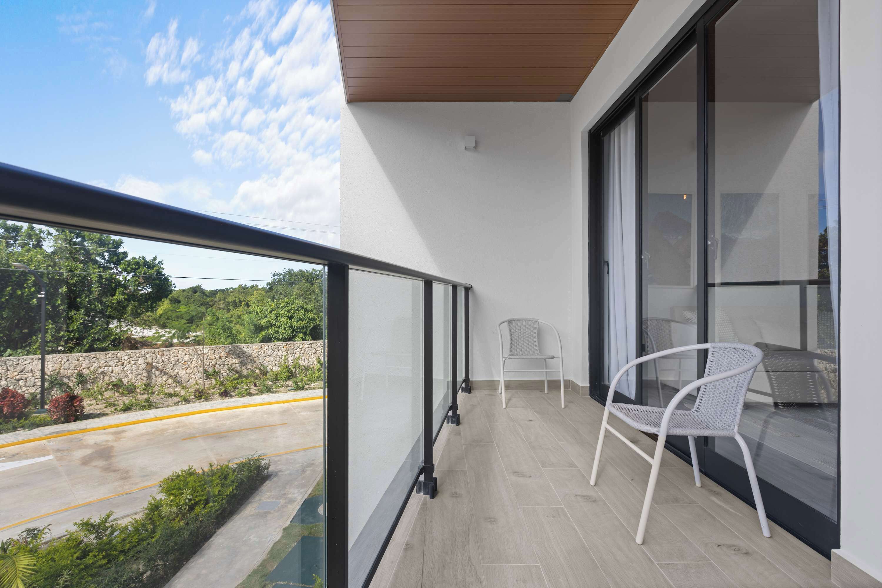 A balcony with a glass railing, a white chair, and a view of a road and greenery.