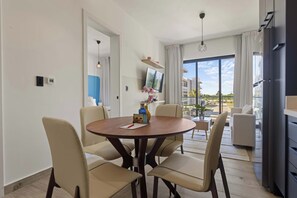 Ein modernes Wohnzimmer mit einem runden Esstisch, beigen Stühlen, einem Flachbildfernseher und einem großen Fenster mit Blick auf Gebäude.