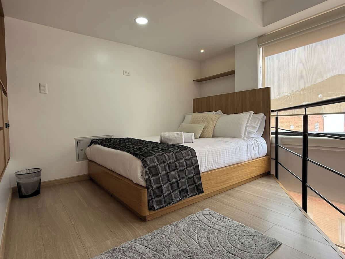 A modern bedroom with a wooden bed, a small bedside table, a trash bin, and a view of the outside through a window.