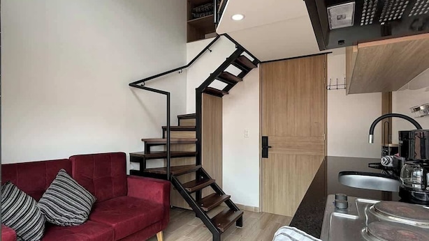 A compact living space with a red sofa, a wooden staircase, a kitchen area, and a small dining table. - NOK Beautiful Apartment in Chico (Bogotá)